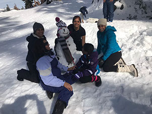 A group of young adults and teens play in the snow, building a snowman.